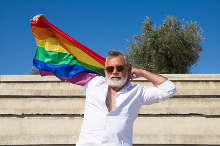 Handsome mature man with beard and grey hair is sitting on the bleachers of an open-air auditorium while holding the gay pride flag in his hands. Concept old age, equality, homosexual, gay, rights.の写真素材