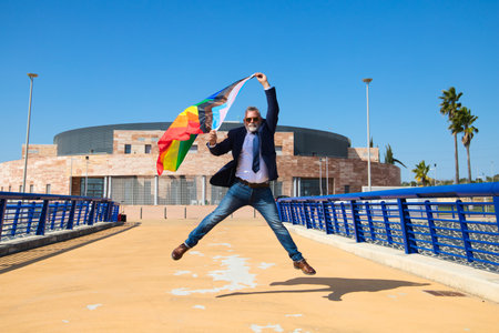 Mature gay man, executive, grey hair, beard, sunglasses, jacket and tie, jumping and waving gay pride flag in the wind. Concept of mature gay man, lgtb pride, equality, rights.の写真素材