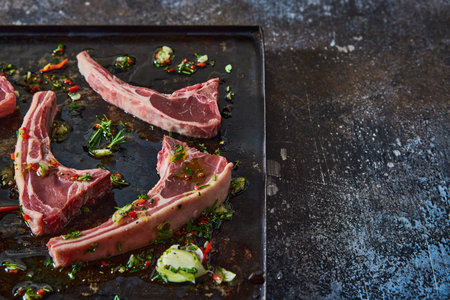 From above of uncooked lamb chops placed on black tray with chopped greens and oil on dark table in light kitchenの写真素材