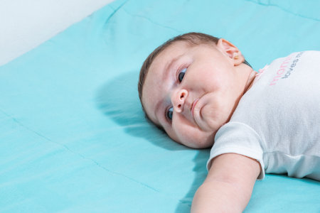 close-up of a beautiful latin baby girl, lying on a bed with blue sheets, pouting while looking at...