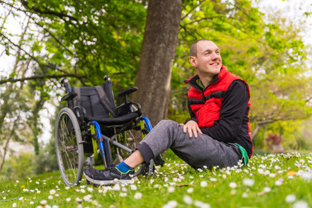 Portrait of a paralyzed young man sitting on the grass next to the wheelchair a daisy flowers, in the spring in a natural parkの写真素材
