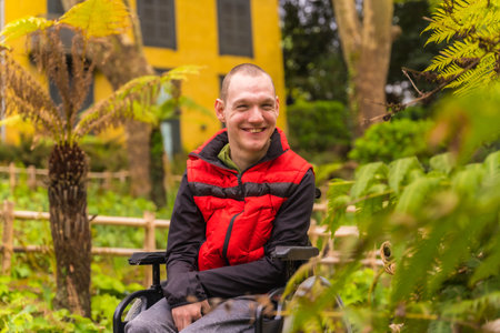 Portrait of a paralyzed young man in a red vest in a public park in the city. Sitting in the wheelchair smilingの写真素材
