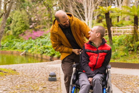 Paralyzed young man in the wheelchair being pushed by a friend in a public city park, having fun in springの写真素材