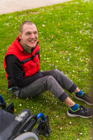 A paralyzed young man sitting on the grass next to the wheelchair next to a tree looking at the cameraの写真素材