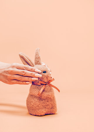 A creative and unusual Easter concept. Female hands hide the eyes of a small puffy brown Easter bunny. Realistic aesthetic look. Contemporary style. Blazing Orange background, soft shadows.の写真素材