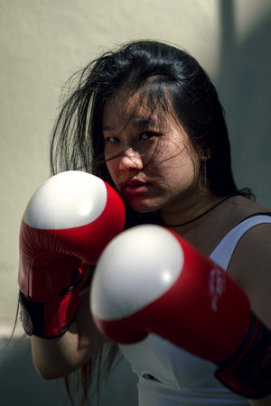 Chinese woman with tough look training with red and white kick boxing gloves looking at the camera.Outdoor sport concept.の写真素材