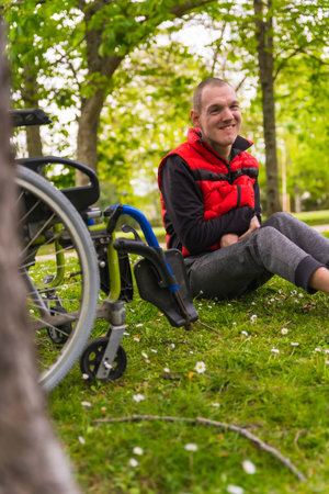 A paralyzed young man sitting on the grass next to the wheelchair next to a tree looking at the cameraの写真素材