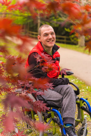 Portrait of a paralyzed young man in a red vest in a public park in the city. Sitting in the wheelchair next to autumn leavesの写真素材