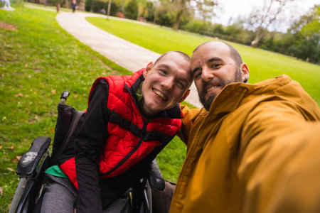 Selfie of a paralyzed young man in a wheelchair with a friend on a bench in a public park in the city, talking and laughingの写真素材