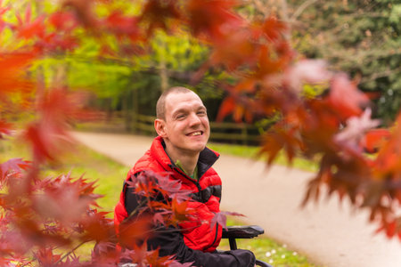 Portrait of a paralyzed young man in a red vest in a public park in the city. Sitting in the wheelchair next to autumn leavesの写真素材