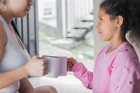 beautiful latin girl with brown skin, sitting on the floor next to her mother toasting with white mugs. the girl looks very happy to her mother while smiling. mother's day celebration.の写真素材