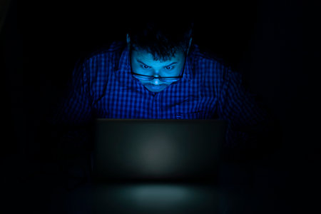 young man typing on laptop keyboard at night and looking over the eyeglass