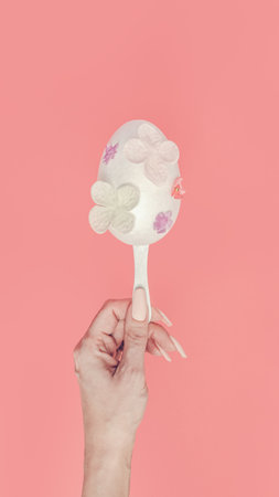 Modern flat lay Easter concept. A female hand holds a white egg on a stick. Decoration with pastel flowers of different sizes. Handmade craft. Spring and romantic concept on nude pastel background.の写真素材
