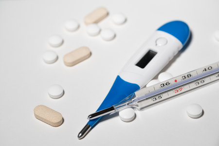 Analog and Digital thermometer on white surface with medicine and pills.