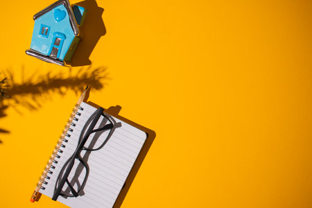 A top view of a neon orange shadowy surface with a notebook, glasses and a house toy on itの写真素材