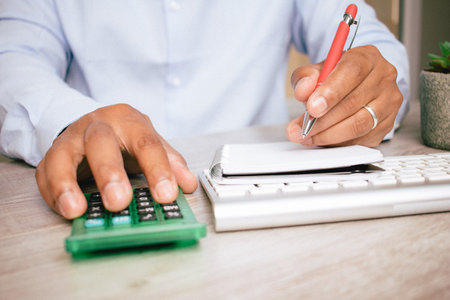 A closeup of a Hispanic man's hands as he uses a calculator and writes in a notebook in his office