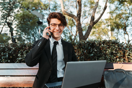 A cheerful professional young Caucasian guy sitting on a bench using his laptop talking on the phoneの写真素材