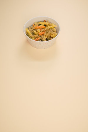 A vertical shot of uncooked tri-color rotini pasta on a bowl isolated on light brown backgroundの写真素材