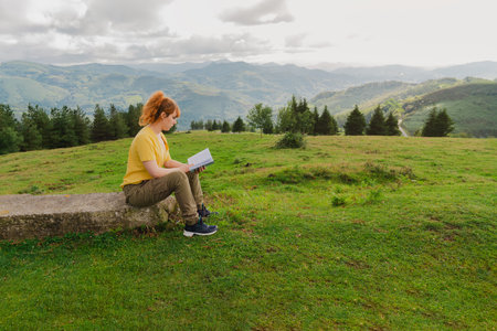 A young female sitting on the stone reading a book in the green field on the background of mountainsの写真素材