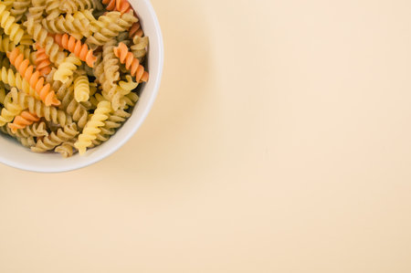A top view of uncooked tri-color rotini pasta on a bowl isolated on light brown backgroundの写真素材