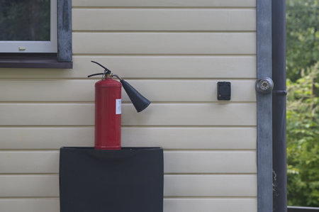A red powder fire extinguisher stands against a wall of white siding. The safetyの写真素材