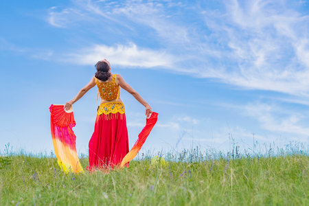 Young woman in bright national dress and fiery fans in her hands is dancing oriental dance against background of blue cloudy sky. Stands his back.の写真素材
