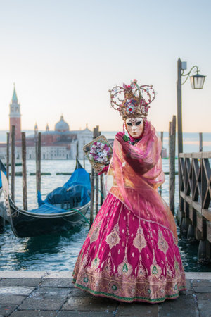 A female wearing a mask and pink carnival costume standing by the water in Venice, Italyの写真素材