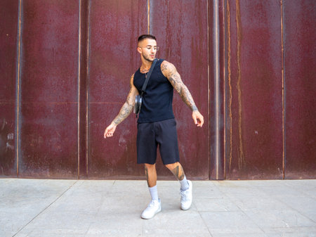 An attractive cool Spanish man with tattoos wearing a tank top with shorts and walking on the...