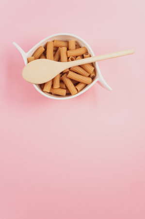 A vertical shot of a bowl of uncooked pasta and a wooden spoon on a pink surfaceの写真素材