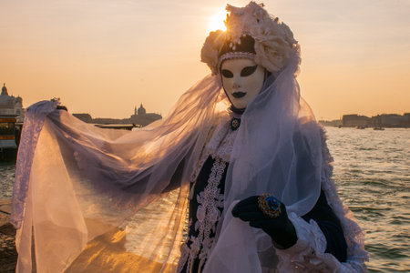 A female wearing a mask and carnival costume standing by the water at sunset in Venice, Italyの写真素材