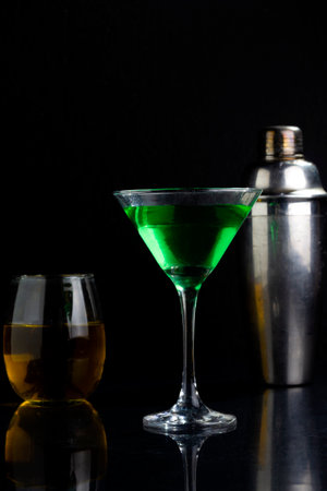 A vertical shot of a vibrant green drink on a dark black background with a shaker and glass behindの写真素材