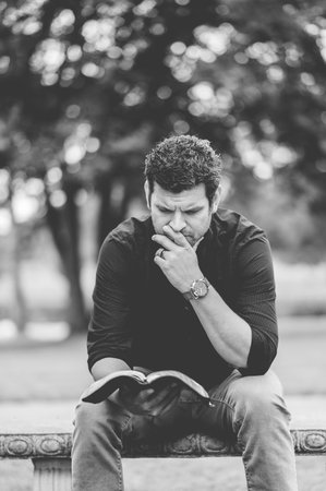 A grayscale of a man reading a book while sitting on the benchの写真素材