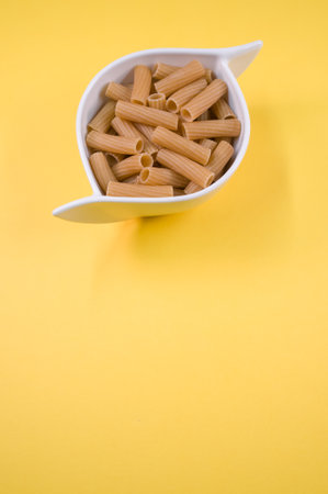 A vertical top view shot of rigatoni in a white bowl on a yellow surface with free space for textの写真素材