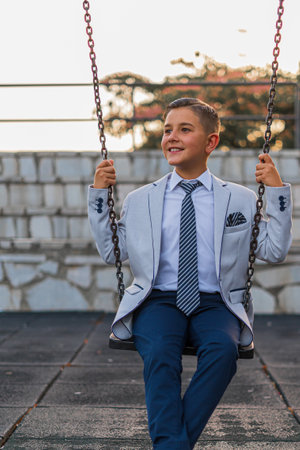 A vertical shot of an adorable male child in a formal suit outfit posing on a swingの写真素材