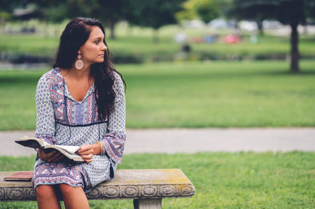 A female taking a pause from reading a bible at a park with grass and treesの写真素材