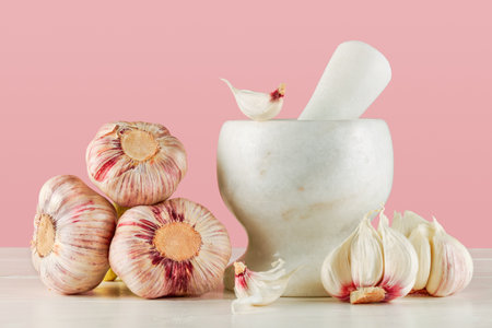 A closeup of fresh garlic and a mortar and pestle on a white wooden table, with a pink backgroundの写真素材