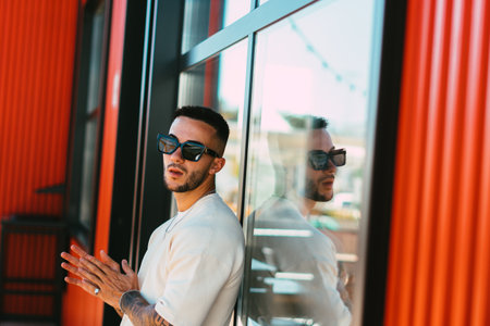 A view of a stylish male wearing sunglasses and posing near the glass wallの写真素材