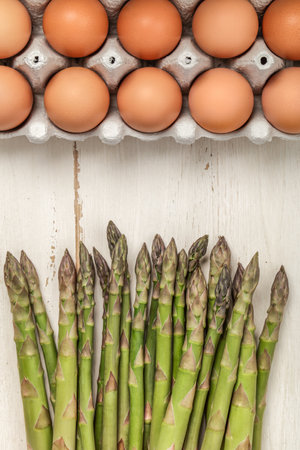 A vertical top view of asparagus and brown eggs in a crate, on a white wooden table