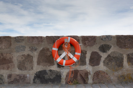 Orange life ring with leash is attached to brick wall in front of blue sky with clouds.の写真素材