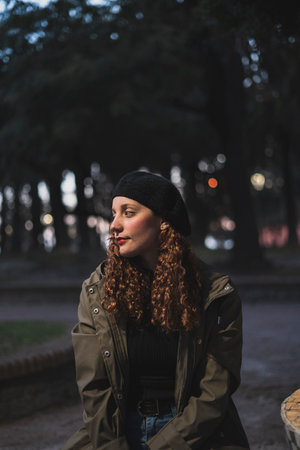 A vertical shot of a female wearing a khaki jacket and a black warm hat sitting in the parkの写真素材
