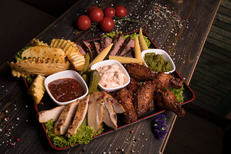 A meat assortment of fried chicken wings, beef, and pork decorated with lettuce, cherry tomatoes, and picklesの写真素材