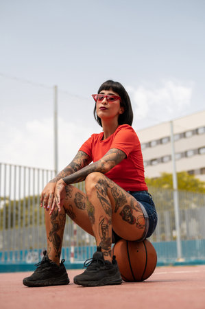 A portrait of a sexy confident Spanish woman with tattoos sitting on a ball at the basketball courtの写真素材