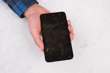 A closeup shot of a person's hand holding a cracked smartphone against a rusty wall backgroundの写真素材