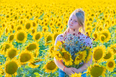 Bright sunny day. Young girl with large bouquet of sunflowers and wildflowers stands in middle of yellow field. Looks into camera. Outdoor recreation.の写真素材