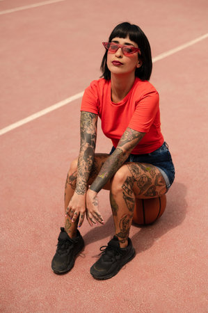 A portrait of a sexy confident Spanish woman with tattoos sitting on a ball at the basketball courtの写真素材
