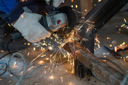 The cut off male hands of a worker use a grinder to clean a weld seam on a metal structure. Bright...