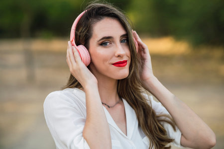 A portrait of a Spanish woman in a white shirt listening to music with pink headphones in a parkの写真素材