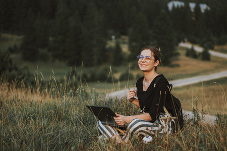 A shallow focus shot of a Bosnian Caucasian woman with a laptop in her lap outdoors in a field,...