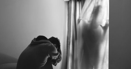 A horizon shot of a scared man sitting on bed and feel afraid from a ghost behind the curtainsの写真素材