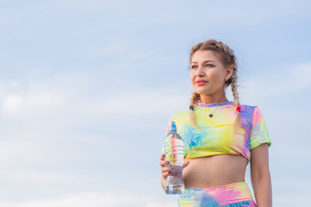 Young beautiful girl in bright tracksuit stands against background of blue summer sky. He holds plastic bottle with drinking water in his hand.の写真素材